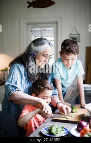 Grand-mère assistant garçon coupant concombre à bord dans la cuisine à accueil Banque D'Images