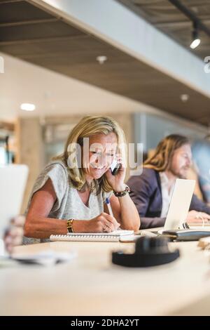 Femme d'affaires souriante écrivant dans un journal tout en parlant sur un smartphone au bureau Banque D'Images