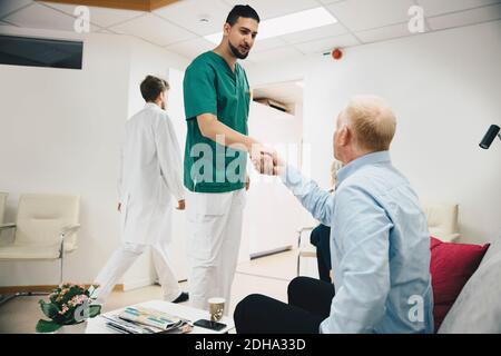 Jeune infirmière de sexe masculin se secouant les mains avec un patient mature assis hall d'entrée à l'hôpital Banque D'Images