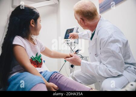 Médecin mature pointant vers un moniteur d'ordinateur pour fille pendant le pouls test à l'hôpital Banque D'Images