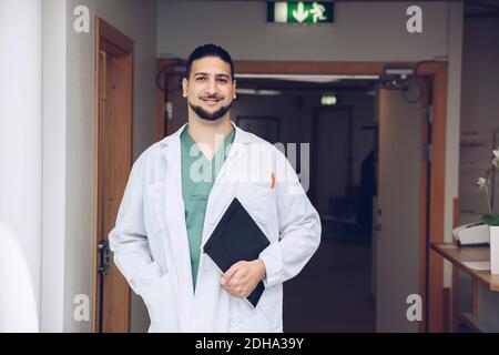 Portrait de jeune homme souriant médecin tenant le bloc-notes tout en étant debout dans le couloir de l'hôpital Banque D'Images