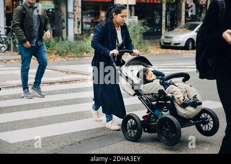 Pleine longueur de mère avec son fils assis dans la poussette croisant route en ville Banque D'Images