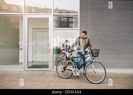 Un père souriant qui fait du vélo avec son fils assis sur le siège de sécurité sur le trottoir de la ville Banque D'Images