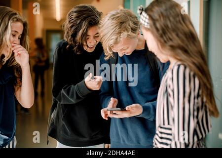 Souriant des élèves de sexe masculin à l'aide d'un smartphone tout en restant debout près d'une femme amis dans le couloir scolaire Banque D'Images