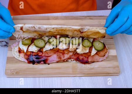 Chef préparant un délicieux sandwich sous-marin à la baguette fraîche. Gros plan des mains dans des gants fermant la baguette sur les tranches de cornichon. Banque D'Images