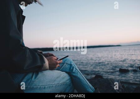 Section médiane d'une femme utilisant un téléphone mobile tout en étant assise sur le bord du lac Banque D'Images