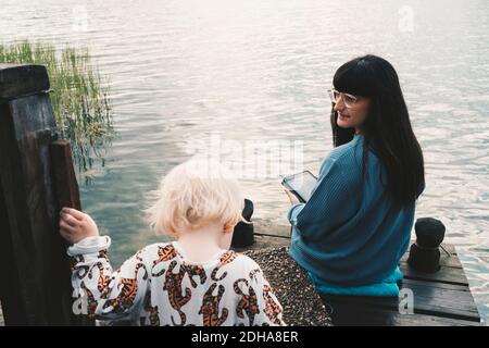 Mère regardant la fille tout en tenant une tablette numérique et assis sur la jetée au-dessus du lac Banque D'Images