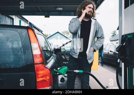 Jeune homme parlant sur le téléphone mobile tout en faisant le plein de voiture à station-service Banque D'Images