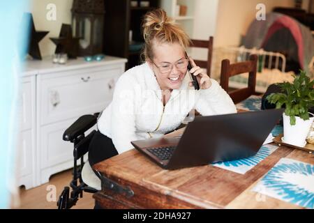 Femme handicapée souriante parlant sur un téléphone portable tout en regardant ordinateur portable dans la maison Banque D'Images