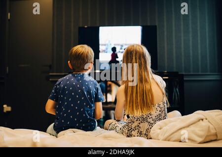 Vue arrière des frères et sœurs regardant la télévision tout en étant assis sur le lit dans la chambre d'hôtel Banque D'Images