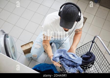 Vue en grand angle du jeune homme qui met des vêtements dans le panier tout en écoutant de la musique Banque D'Images