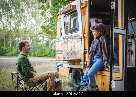 Femme souriante se penchée sur la porte de la caravane tout en parlant avec un homme ami assis pendant le camping en forêt Banque D'Images