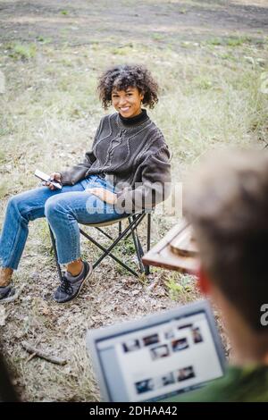 Jeune femme souriante assise sur une chaise tout en regardant un homme ami pendant le camping en forêt Banque D'Images