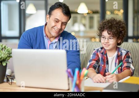 Portrait d'un père hispanique d'âge moyen et joyeux qui passe du temps avec son fils. Petit garçon assis au bureau avec son père et faisant ses devoirs, à l'aide d'un ordinateur portable. Étude en ligne, concept de famille Banque D'Images