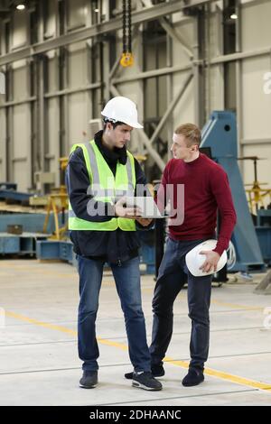 Jeunes hommes, ingénieur d'usine et chef ingénieur dans une robe de travail et casque blanc tenant un smartphone et un ordinateur portable organisant et contrôlant le processus de travail Banque D'Images