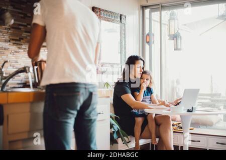 La mère travaille sur un ordinateur portable tandis que la fille regarde le père debout dans la cuisine Banque D'Images