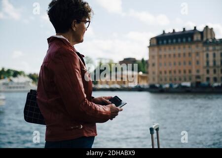 Vue latérale d'une femme âgée tenant un téléphone portable debout par mer en ville Banque D'Images