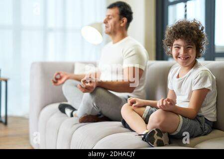 Joyeux garçon d'école latine souriant à la caméra tout en méditant avec son père sur un canapé à la maison. Concept de saines habitudes de vie Banque D'Images