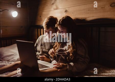 Sourire à l'aide d'un ordinateur portable tout en étant assis sur le lit contre le mur à la maison Banque D'Images