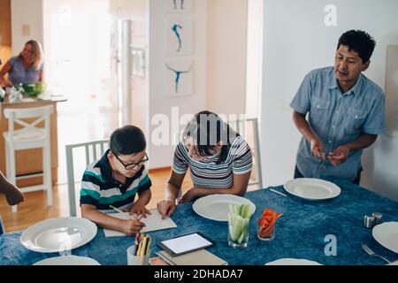 Adolescente aidant le garçon à faire ses devoirs pendant que son père et sa sœur debout à la table Banque D'Images