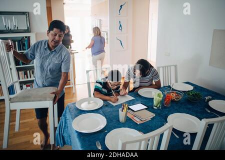 Adolescente aidant frère dans les devoirs pendant que le père arranger des chaises à la table à manger Banque D'Images