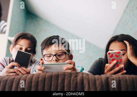 Vue en contre-plongée d'un garçon regardant une tablette numérique pendant couché avec des sœurs utilisant un téléphone portable sur le lit à la maison Banque D'Images