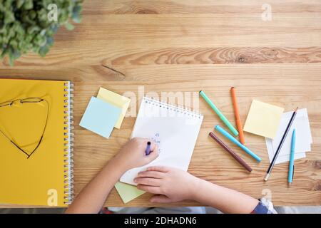 Vue de dessus des mains d'un garçon d'école latino-américain en train de dessiner sur le papier à l'aide de crayons tout en étudiant à la maison. Concept de homeschooling Banque D'Images