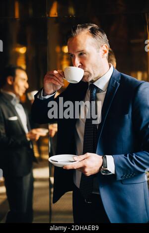 Homme d'affaires mûr buvant du café tout en se tenant contre la réflexion de l'homme collègue sur verre Banque D'Images