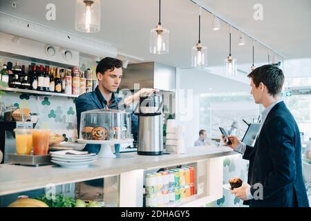 Homme d'affaires tenant un muffin et utilisant le téléphone tout en faisant du café par le propriétaire à la cafétéria du bureau Banque D'Images