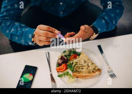 Milieu de section de l'homme d'affaires vérifiant le taux de sucre dans le sang tout en ayant de la nourriture à la table Banque D'Images