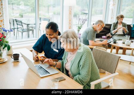 Femme souriante aidant une femme âgée à faire des achats en ligne avec carte de crédit et ordinateur portable dans le cdm de soins infirmiers Banque D'Images