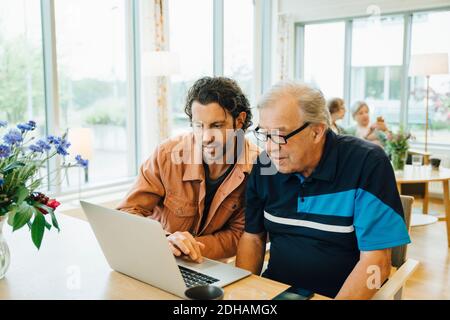 Homme assistant grand-père utilisant un ordinateur portable à la table à manger à la retraite accueil Banque D'Images
