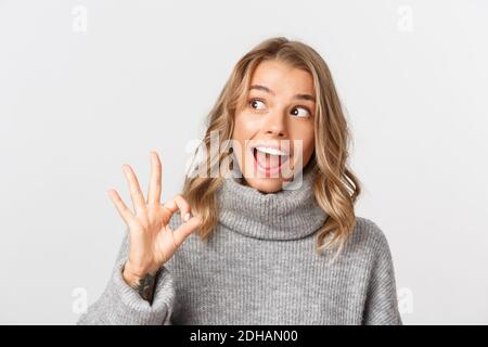 Gros plan de la jeune fille blonde excitée dans un chandail gris, regardant stupéfait à gauche et montrant le signe OK, debout sur fond blanc Banque D'Images