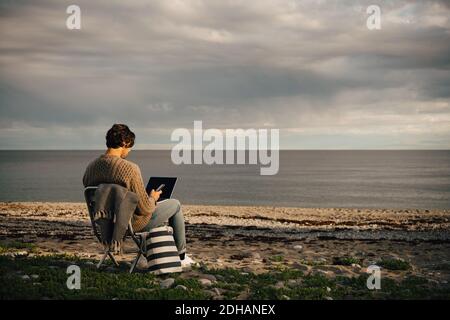 Homme adulte moyen utilisant un ordinateur portable et un téléphone portable assis sur le rivage de la mer à la plage contre le ciel Banque D'Images