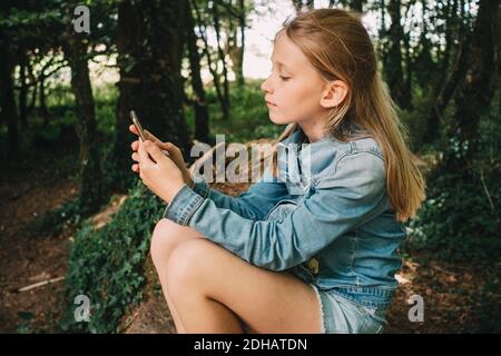 Vue latérale d'une fille blonde à l'aide d'un smartphone en forêt Banque D'Images