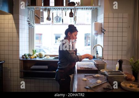 Vue latérale d'un chef mûr parlant sur un téléphone portable pendant préparation de la nourriture dans la cuisine du restaurant Banque D'Images