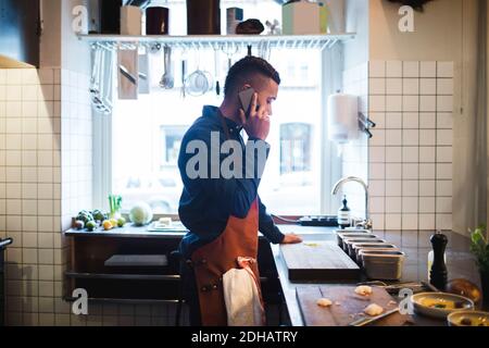 Vue latérale d'un chef mûr parlant sur un téléphone portable pendant au comptoir de la cuisine dans le restaurant Banque D'Images
