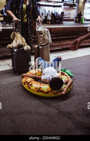 Garçon allongé sur un anneau gonflable tandis que la mère se tient debout avec des bagages à la plate-forme de la gare Banque D'Images