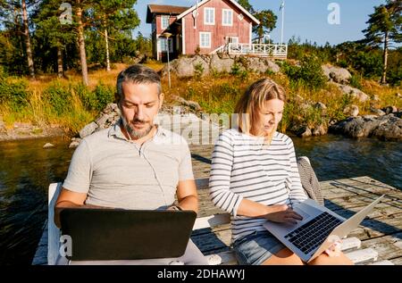 Couple utilisant un ordinateur portable tout en étant assis sur un banc ensemble à la jetée en été Banque D'Images