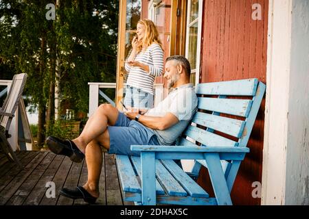 Homme souriant assis sur le banc pendant qu'une femme parle en mobile téléphone sous le porche Banque D'Images