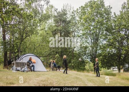 Les enfants jouent au football tandis que la famille s'assoit sous la tente camping Banque D'Images