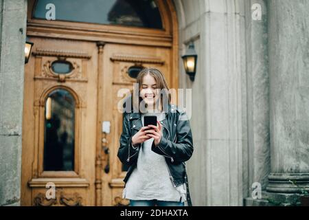 Fille souriante utilisant un téléphone portable tout en se tenant contre le bâtiment Banque D'Images