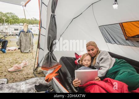 Fille regardant la tablette numérique tout en étant couché avec la mère dans la tente au camping Banque D'Images