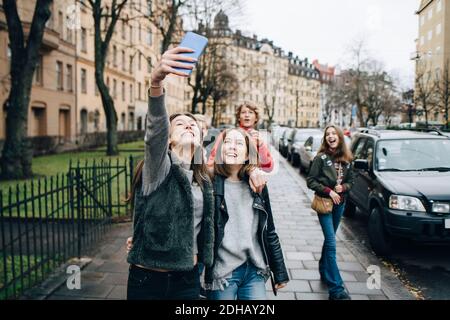 Une fille souriante prend son selfie sur son smartphone avec ses amis pendant que vous l'emperez debout sur le trottoir de la ville Banque D'Images