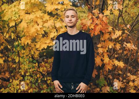 Adolescent confiant avec des cheveux blonds debout contre des érables en automne Banque D'Images