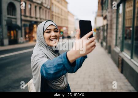 Jeune femme musulmane gaie portant le hijab emportant le selfie sur le trottoir en ville Banque D'Images