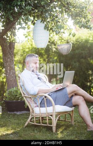 Homme mature qui tape sur un ordinateur portable tout en étant assis sur une chaise en bois dans l'arrière-cour Banque D'Images