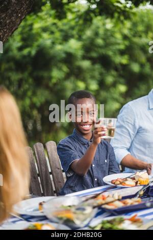 Un garçon souriant tenant un verre tout en étant assis avec sa famille arrière-cour pendant la fête Banque D'Images