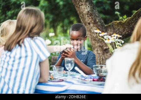 Garçon souriant regardant le téléphone tout en étant assis avec des amis à table à manger dans l'arrière-cour Banque D'Images