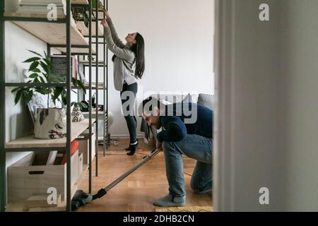 Homme passant l'aspirateur sur le sol pendant qu'une femme nettoie l'étagère à la maison Banque D'Images